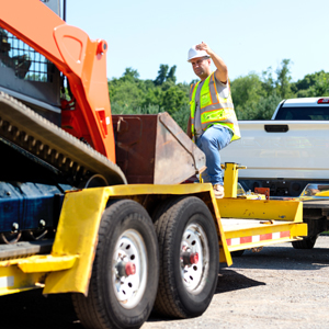 Construction working guiding a skid steer onto a heavy duty trailer connected to a pick up truck
