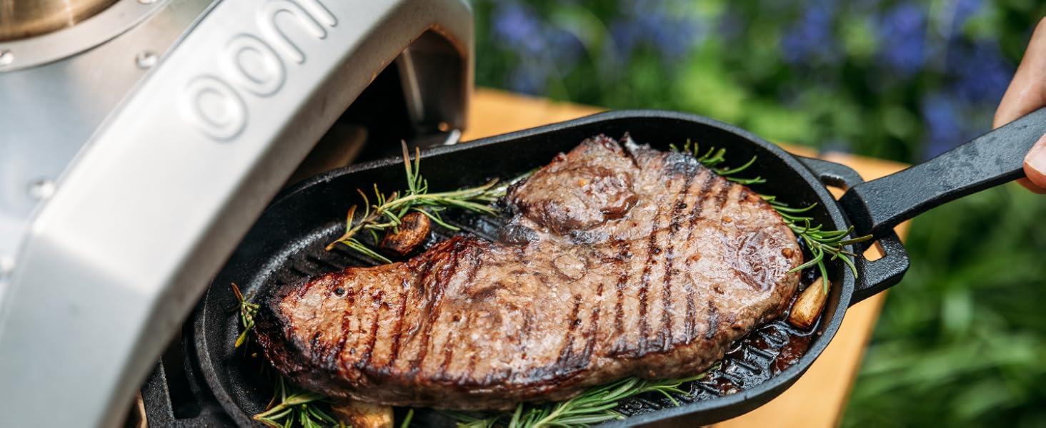 Outdoor grill with 'Ooni' logo cooking a large steak on a cast iron skillet. Herbs visible around the meat.
