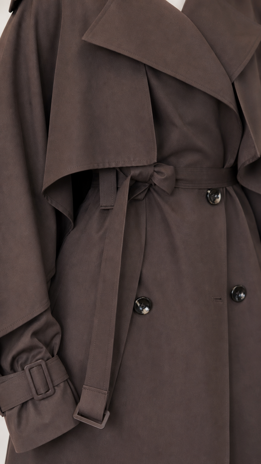 Close-up of dark brown trench coat cape overlay, belt buckle, cuff strap, and buttons