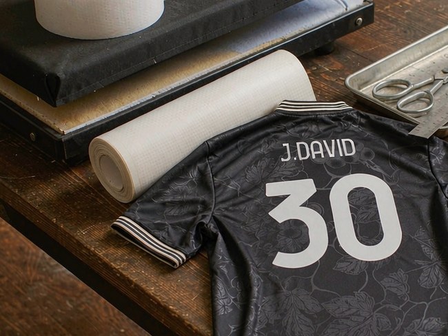 Close-up of a black Juventus third shirt printed J. DAVID 30 lying on a workshop table beside a roll of transfer film