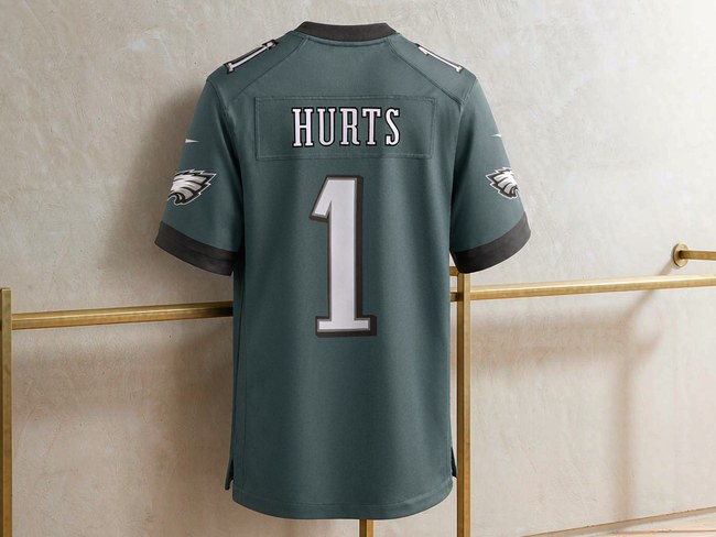 Philadelphia Eagles midnight green home jersey with HURTS 1 printed on the back, hanging on a brass rail against a cream plaster wall