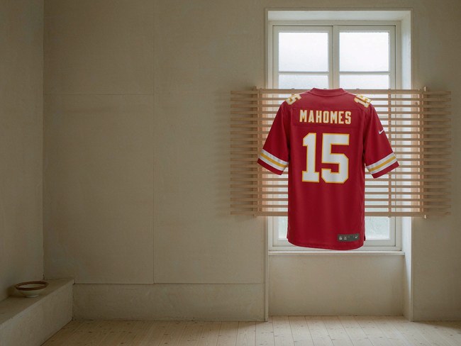 A red Kansas City Chiefs football jersey hanging on a wooden drying rack in front of a bright window, air-drying naturally
