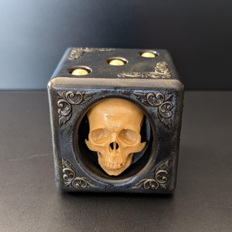 May include: A black cube-shaped die featuring ornate corner scrollwork and three gold-colored circular pips on the top face. The front face displays a circular cutout containing a realistic, beige-colored human skull sculpture centered within.