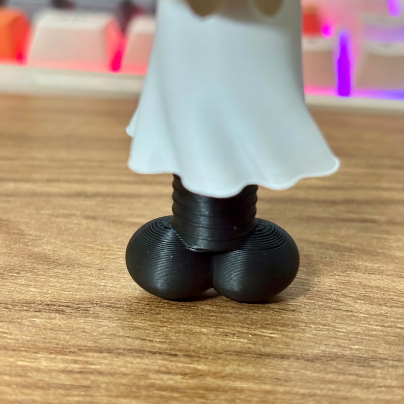 May include: A small figurine featuring a white, flowing, skirt-like garment positioned above a black, cylindrical, ribbed connector piece that attaches to two rounded, black, spherical feet with visible 3D printed layer lines.