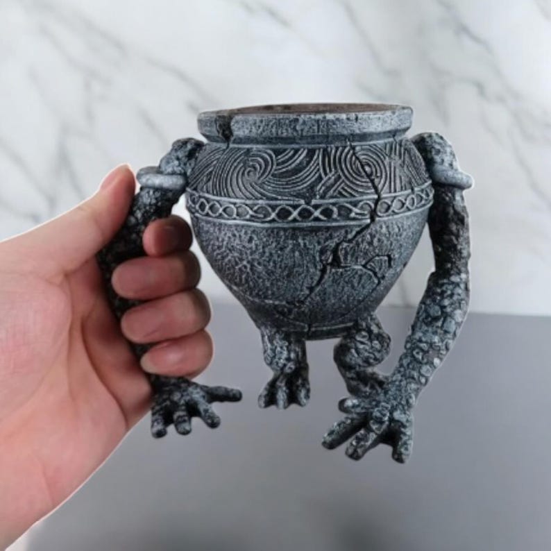 May include: A gray, stone-like planter shaped like a creature with two arms and legs. The planter has a textured surface and a cracked design.