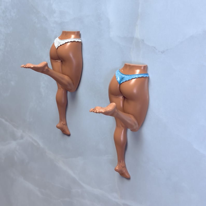 May include: Two wall-mounted sculptures of human legs and buttocks. One sculpture wears white lace underwear, the other wears blue lace underwear. The sculptures are painted with a glossy finish and are mounted on a light gray wall.