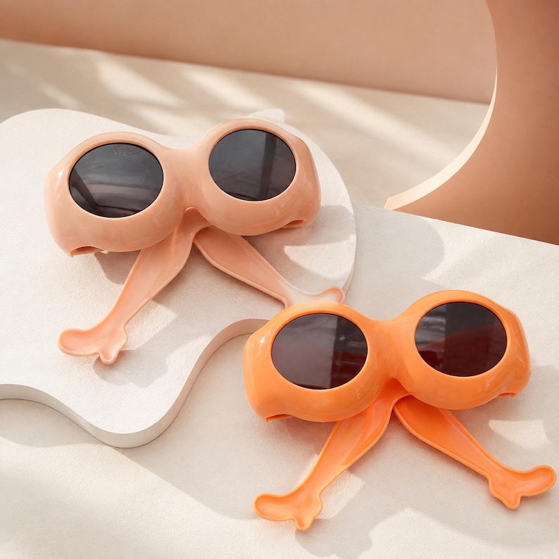 May include: Two pairs of novelty sunglasses featuring thick, rounded frames and integrated leg-shaped temples. One pair is light peach and the other is bright orange. Both pairs have dark, circular lenses and a smooth, matte-finish plastic construction.