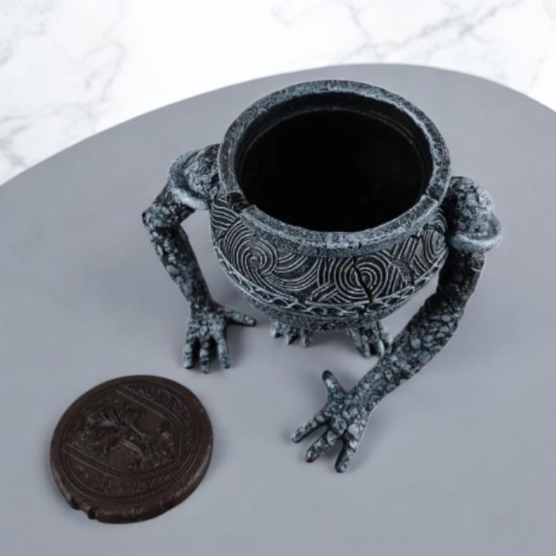 May include: A gray and white ceramic planter shaped like a creature with a cauldron for a head. The planter has detailed carvings and a textured surface.