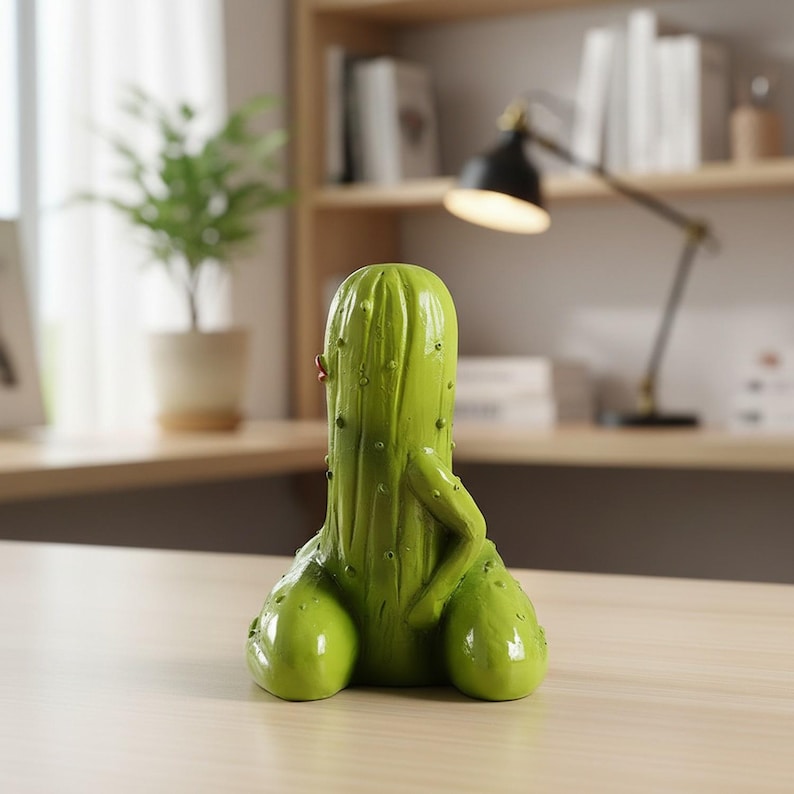 May include: A bright green, anthropomorphic cactus figurine with a smooth, glossy finish. The figure features a central vertical body with small raised bumps, two rounded base sections, and a single arm resting against its side.