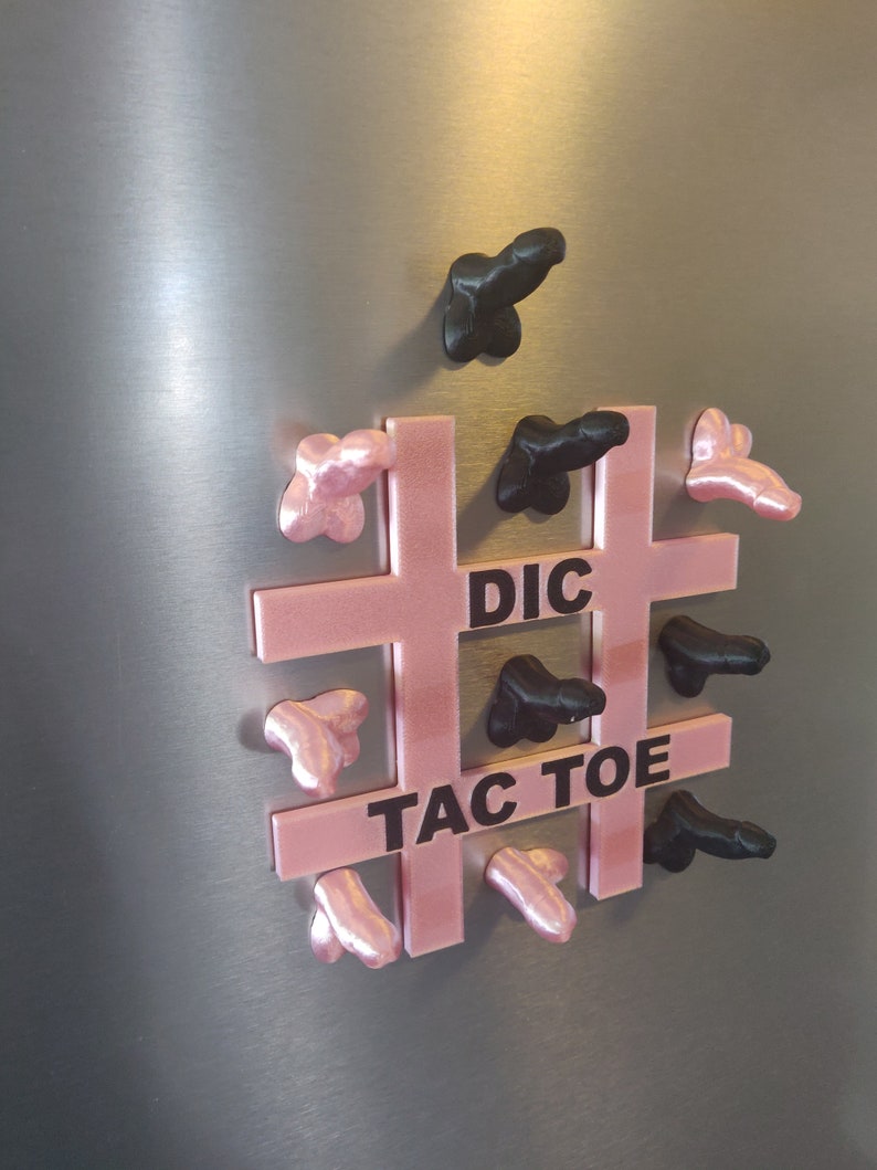 May include: A novelty Tic-Tac-Toe game with a pink grid and black and pink phallic-shaped game pieces. The words "DIC TAC TOE" are printed on the grid. The game is displayed on a metallic surface.