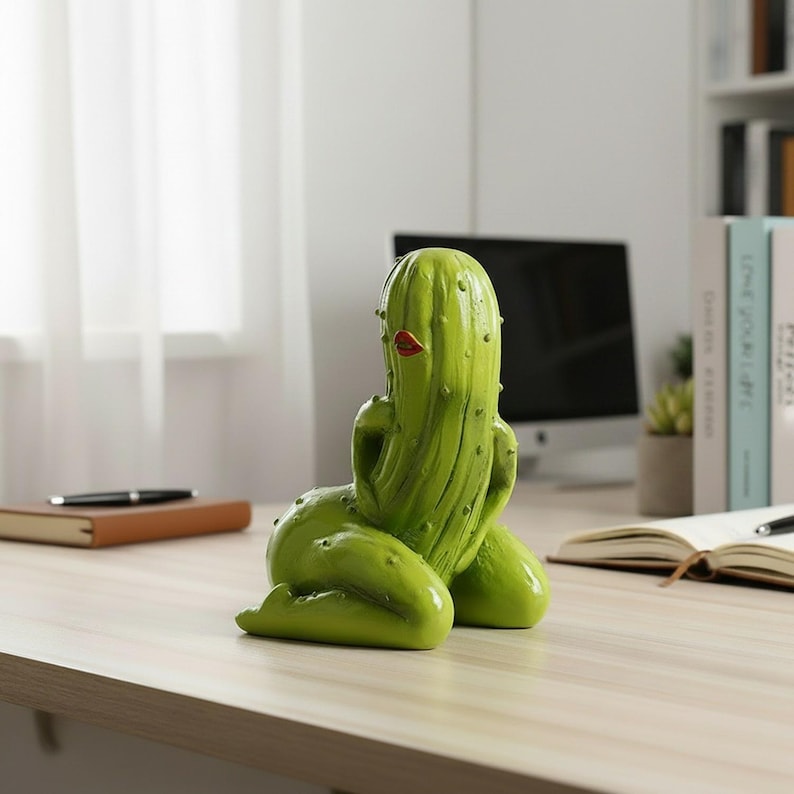 May include: A lime green cactus figurine shaped like a kneeling figure with a smooth, rounded body and small raised bumps. The figure features a pair of bright red lips positioned on the upper portion of the cactus, creating a distinct facial detail.