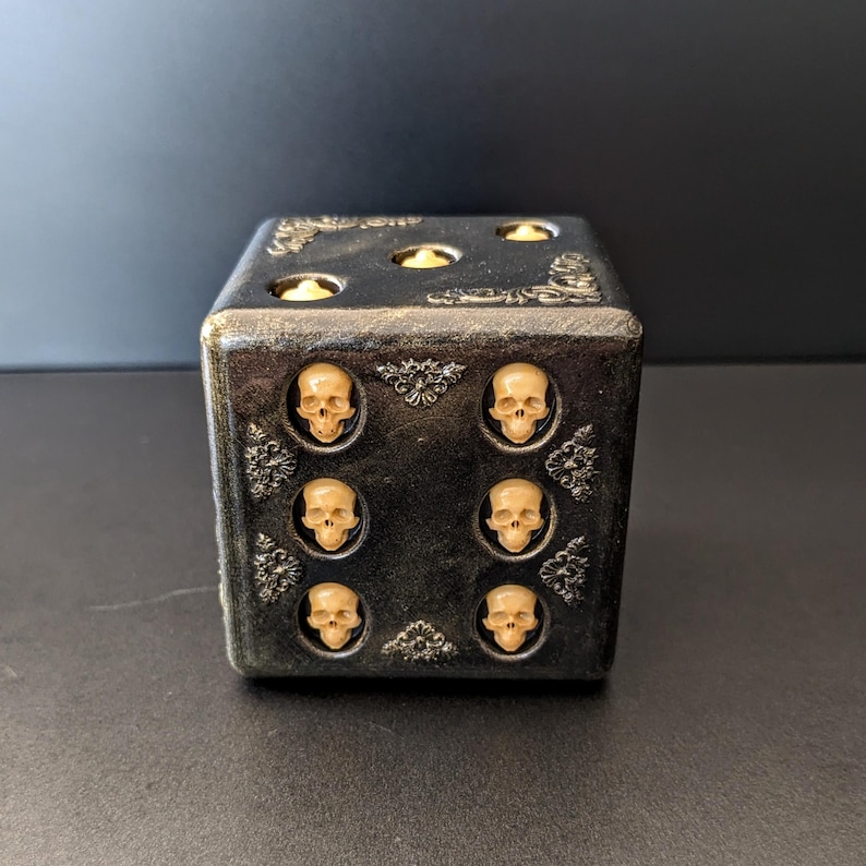 May include: A black cube-shaped die with a distressed finish and ornate filigree corner accents. The top face displays three circular indentations, while the front face features six small, cream-colored skull motifs arranged in two vertical columns.
