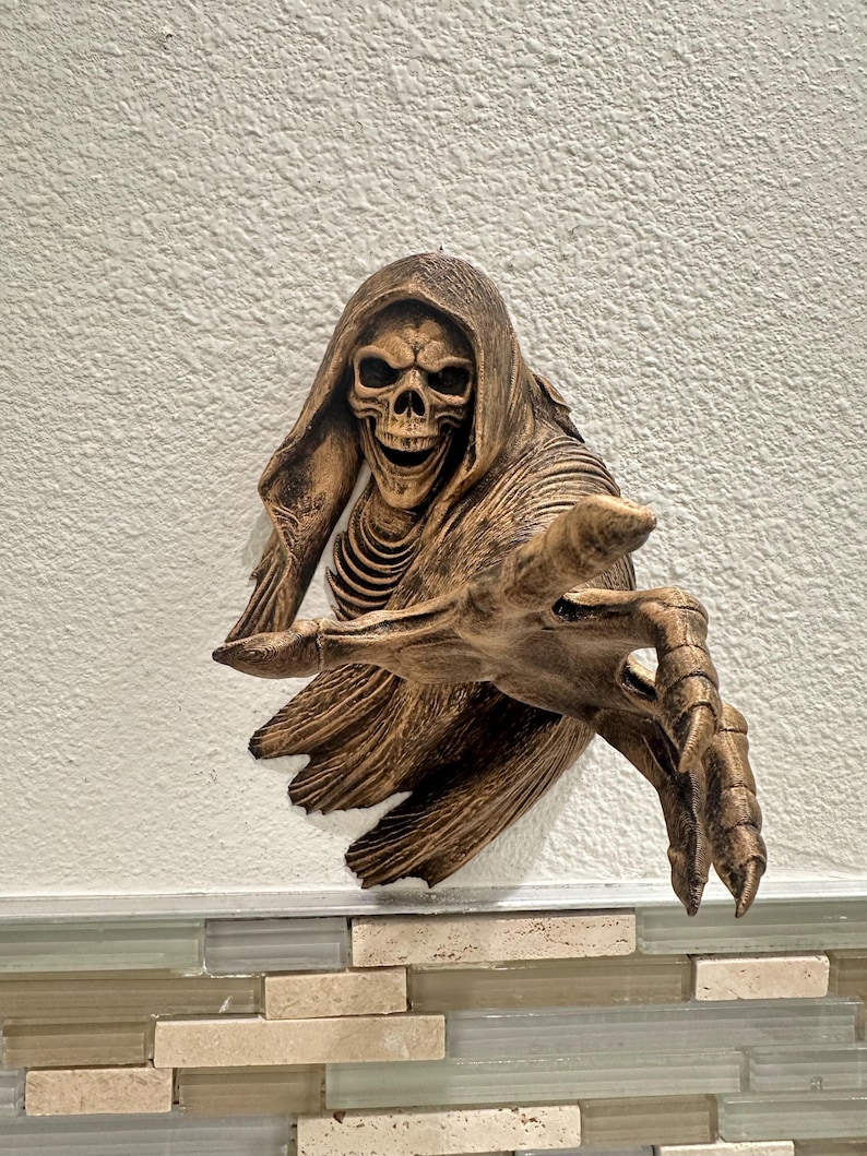 May include: A bronze-colored wall sculpture of a skeletal figure with a hooded cloak and outstretched hand. The figure has a skull face with visible teeth and rib cage. The sculpture is mounted on a textured white wall.