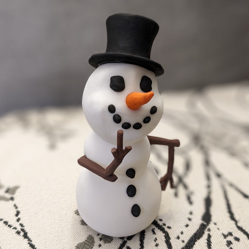 May include: A white snowman figurine with a black top hat and orange carrot nose. The snowman has black button eyes and a black smile.