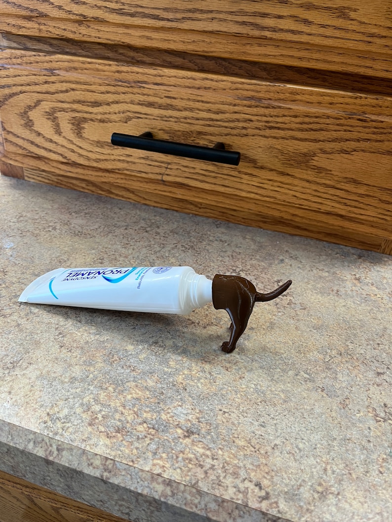May include: A white tube of ProNamel toothpaste with a brown plastic dog-shaped dispenser. The dispenser is shaped like a dachshund and has a long tail.
