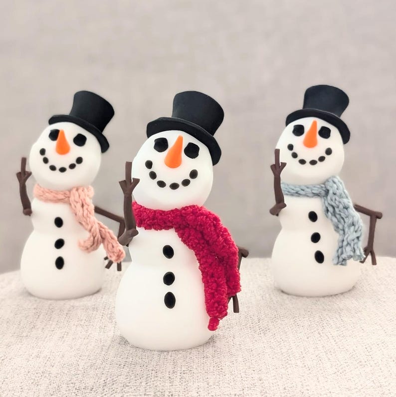 May include: Three white snowmen with black eyes and mouths, wearing black top hats and colorful scarves. The snowman on the left has a pink scarf, the middle snowman has a red scarf, and the snowman on the right has a blue scarf.