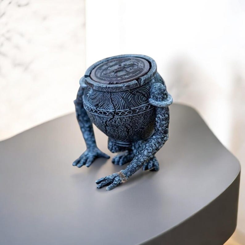 May include: A gray stone-like figurine of a creature with a round body and four legs. The creature is holding a round lid in its hands. The figurine is detailed with intricate carvings and a cracked surface.
