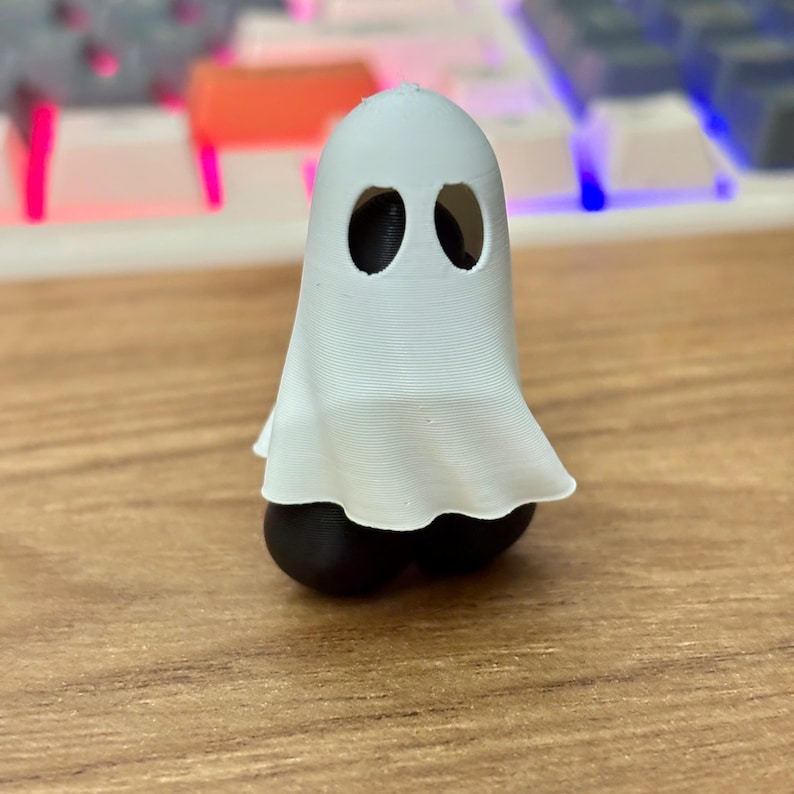 May include: A small 3D printed figurine featuring a white ghost sheet draped over a black base. The ghost has two oval-shaped eye cutouts, and the white material shows fine horizontal layer lines from the printing process, creating a textured surface.