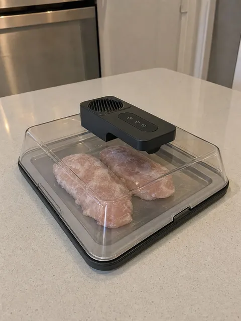 Black defrosting box fully assembled over frozen chicken on a kitchen counter