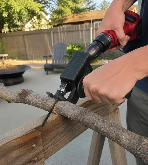 Black drill-powered jigsaw adapter trimming a dry branch on a backyard sawhorse