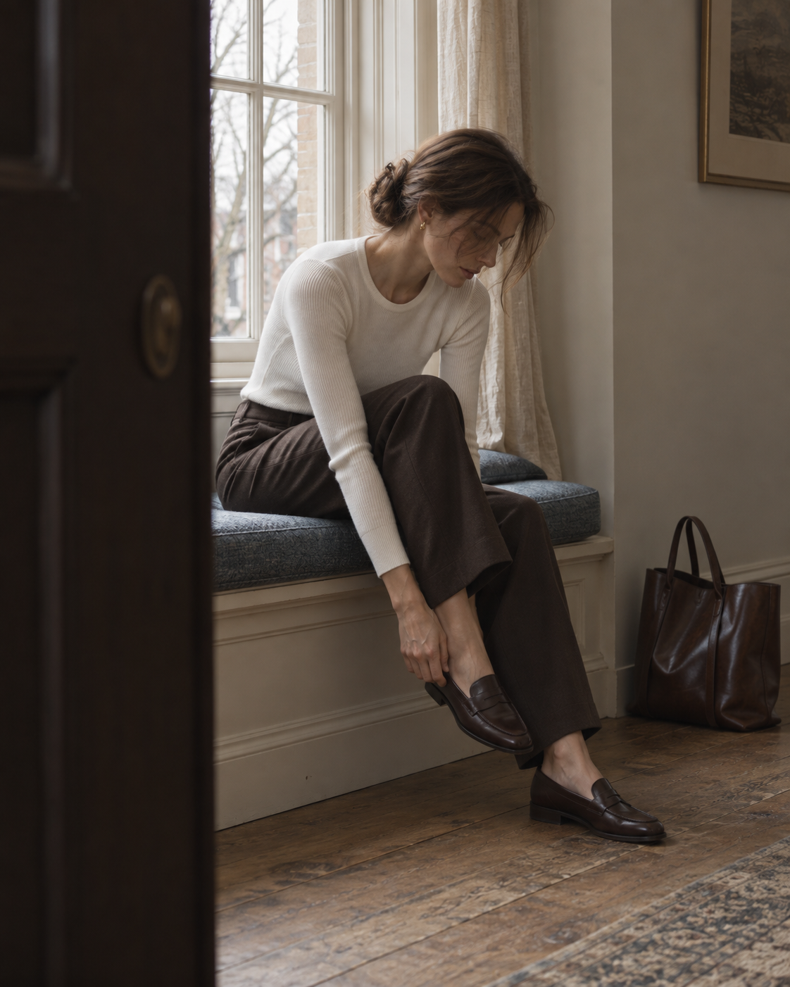 A woman puts on a loafer beside a tall window.