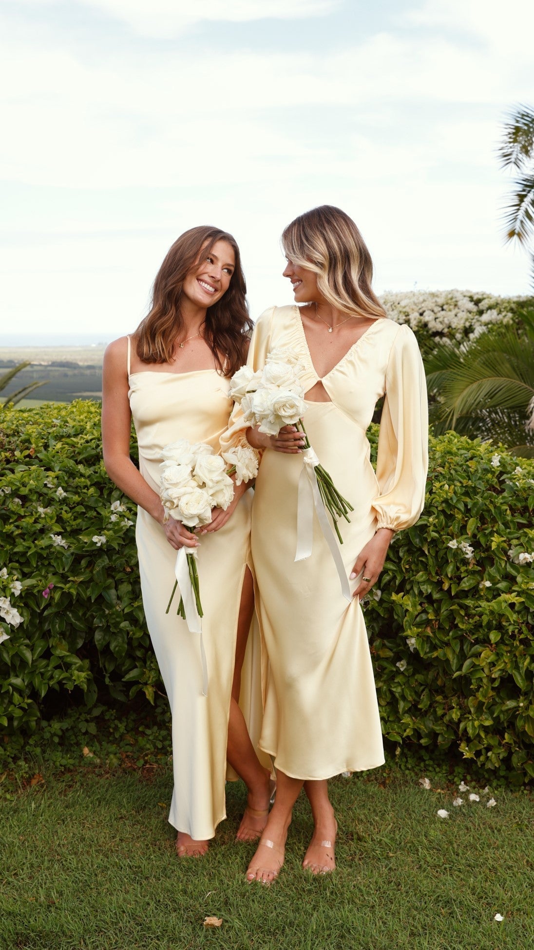 Two women in cream ruched maxi dresses holding white flowers