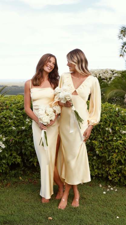 Two women in cream ruched maxi dresses holding white flowers