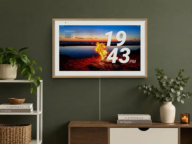 A wall-mounted Digital Calendar displaying a photo of a sunset campfire with a large digital clock and daily schedule, placed above a wooden cabinet with plants and books.