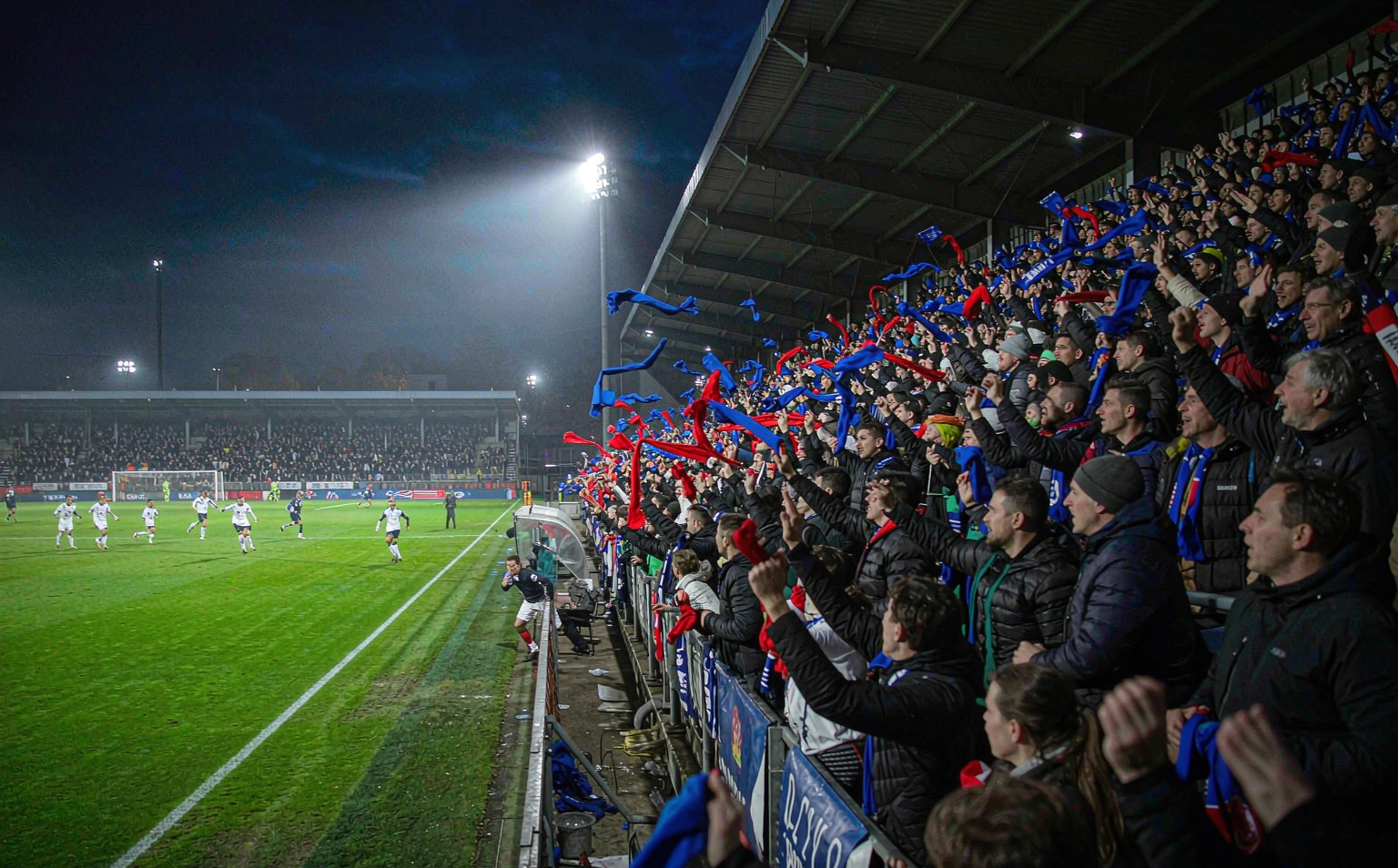Floodlit European football night with crowd holding plain scarves over glowing pitch