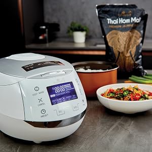 Białe urządzenie kuchenne z wyświetlaczem cyfrowym, prawdopodobnie multi-cooker. Miska kolorowych potraw i składników widoczna w pobliżu.