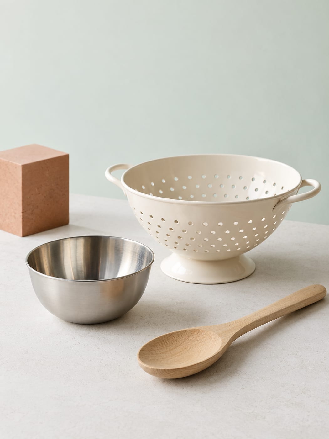 Ivory colander, steel bowl and wood spoon against a pale mint background