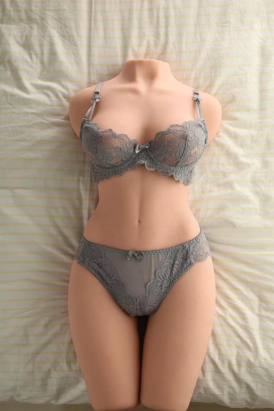 Life-size torso doll in grey lace lingerie lying on a bed with light-colored striped sheets, showcasing realistic body curves and skin texture.