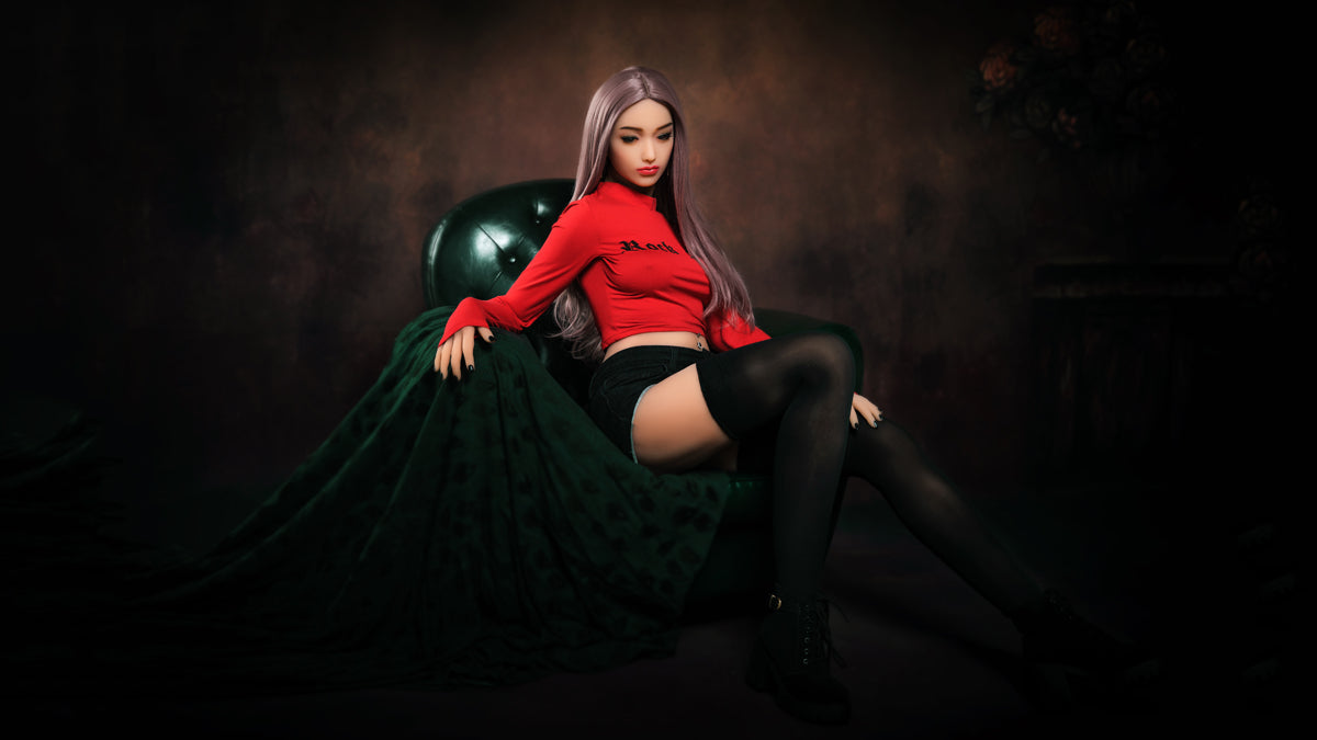 158cm life-size realistic TPE sex doll Anita sitting on a green leather chair, wearing a red crop top, black shorts, black stockings, and black shoes, with long straight hair.