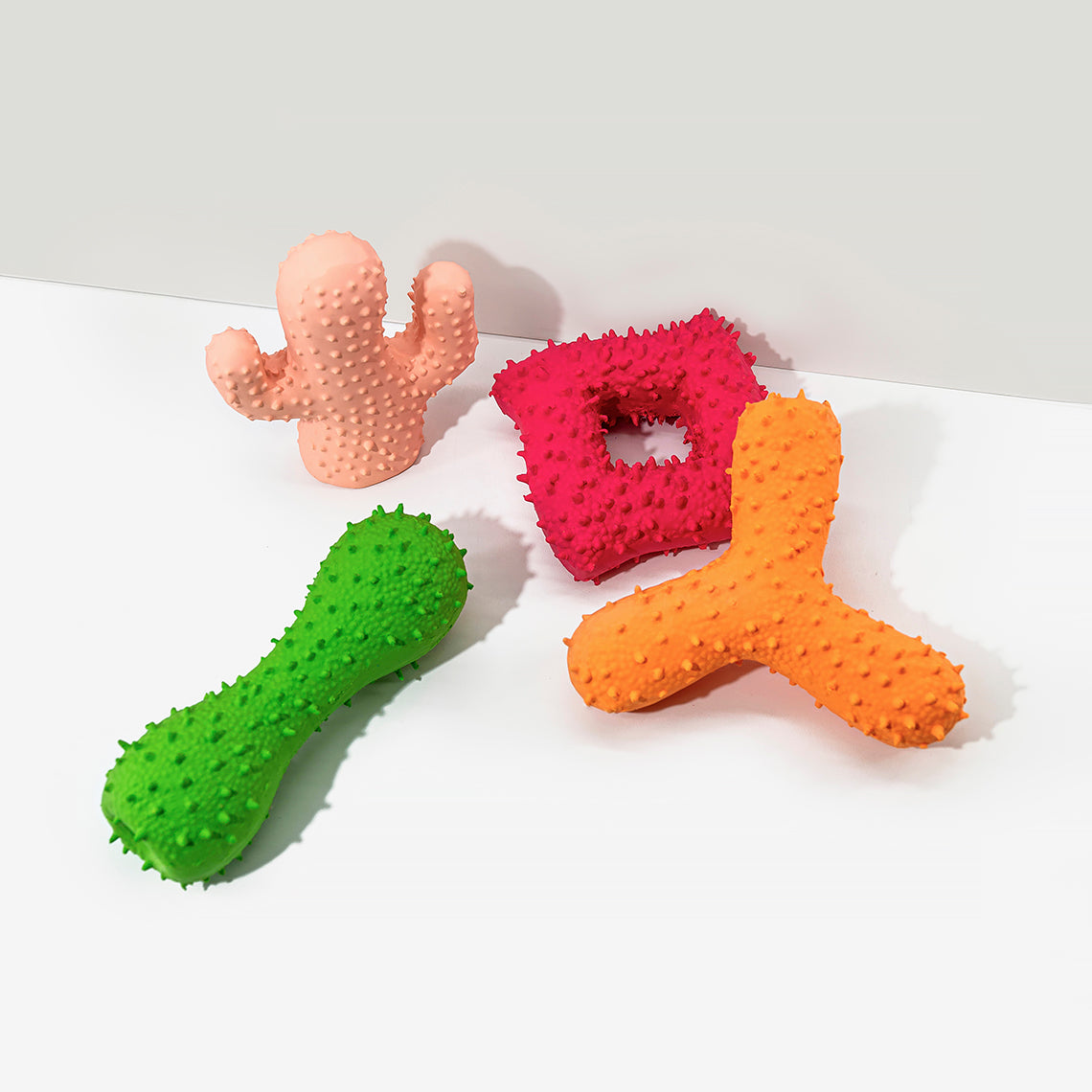 Four dog toys on the white background, include a pink cactus, a red square, a green dumbbell, and an orange boomerang.