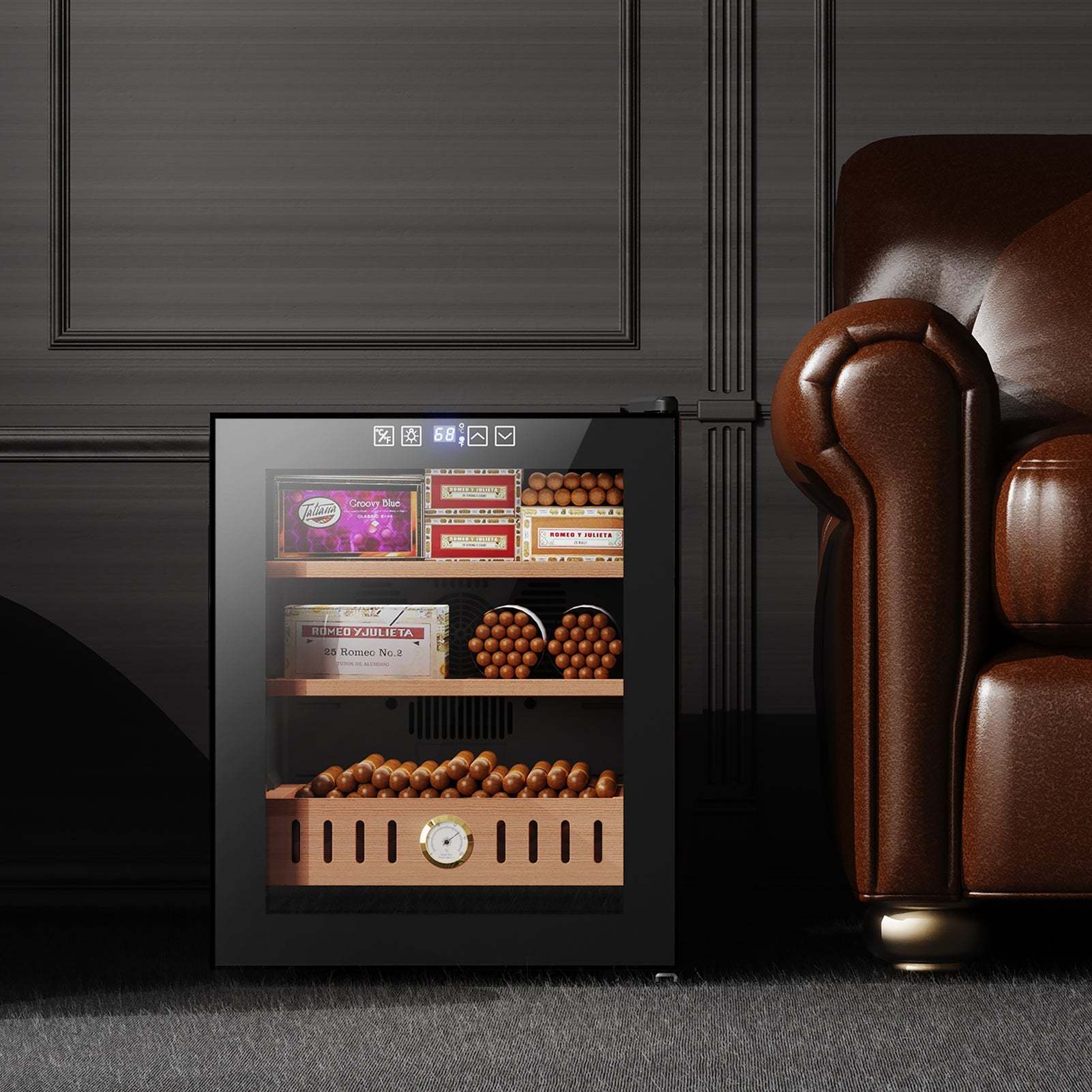 The image shows the Mojgar cigar humidor placed in a stylish home setting next to a brown leather chair. The humidor is neatly organized with cigar boxes and a wooden drawer. The temperature display is visible on the top, indicating a well-maintained storage environment.