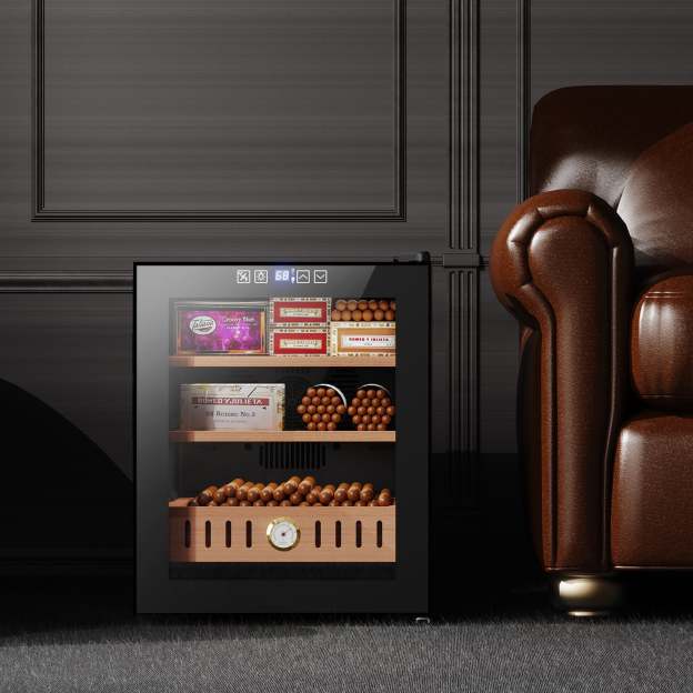 The image shows the Mojgar cigar humidor placed in a stylish home setting next to a brown leather chair. The humidor is neatly organized with cigar boxes and a wooden drawer. The temperature display is visible on the top, indicating a well-maintained storage environment.